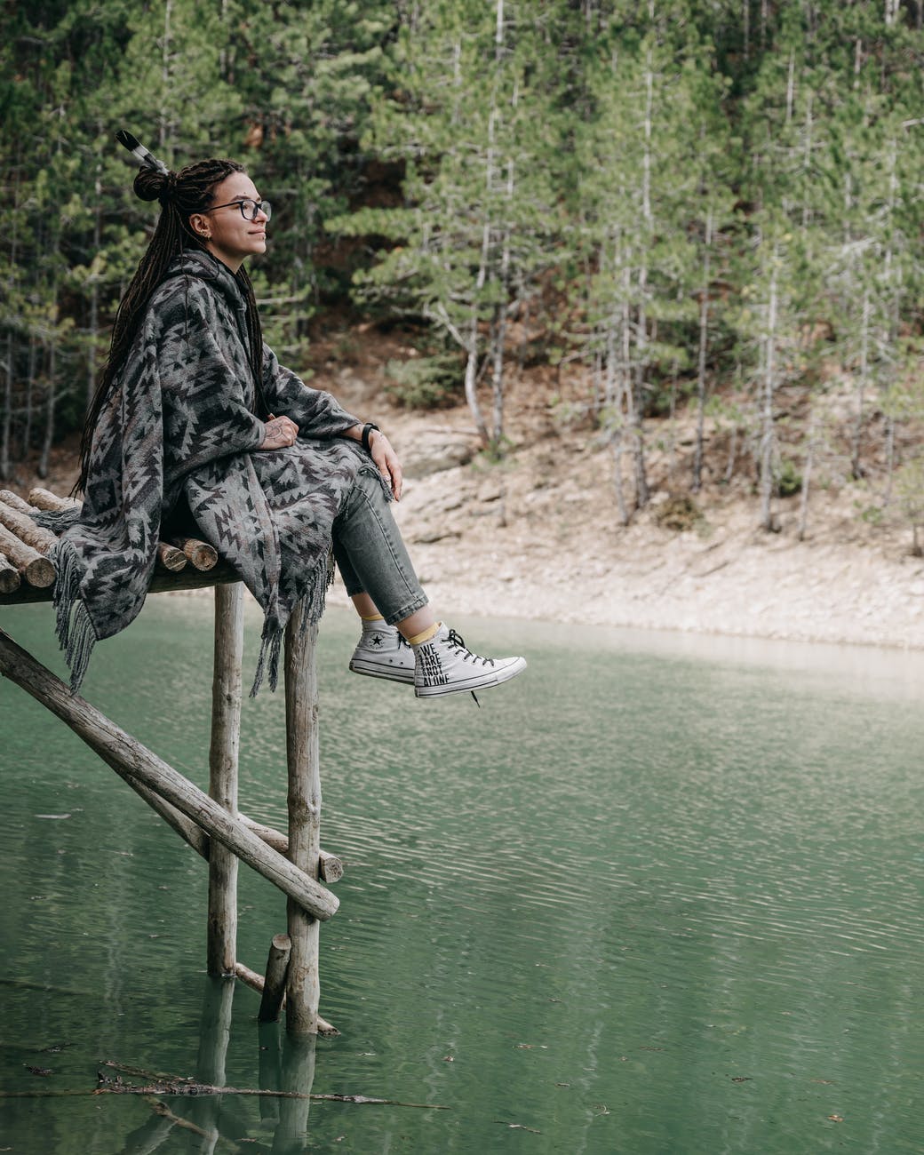 pensive female in poncho sitting on wooden pier above river in forest